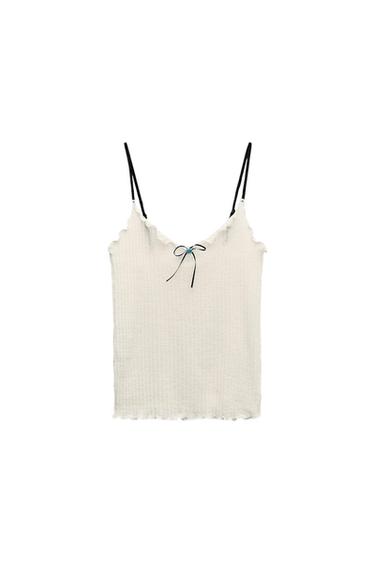 White camisole with thin black straps, wavy trim, and decorative bow at the neckline. White background.