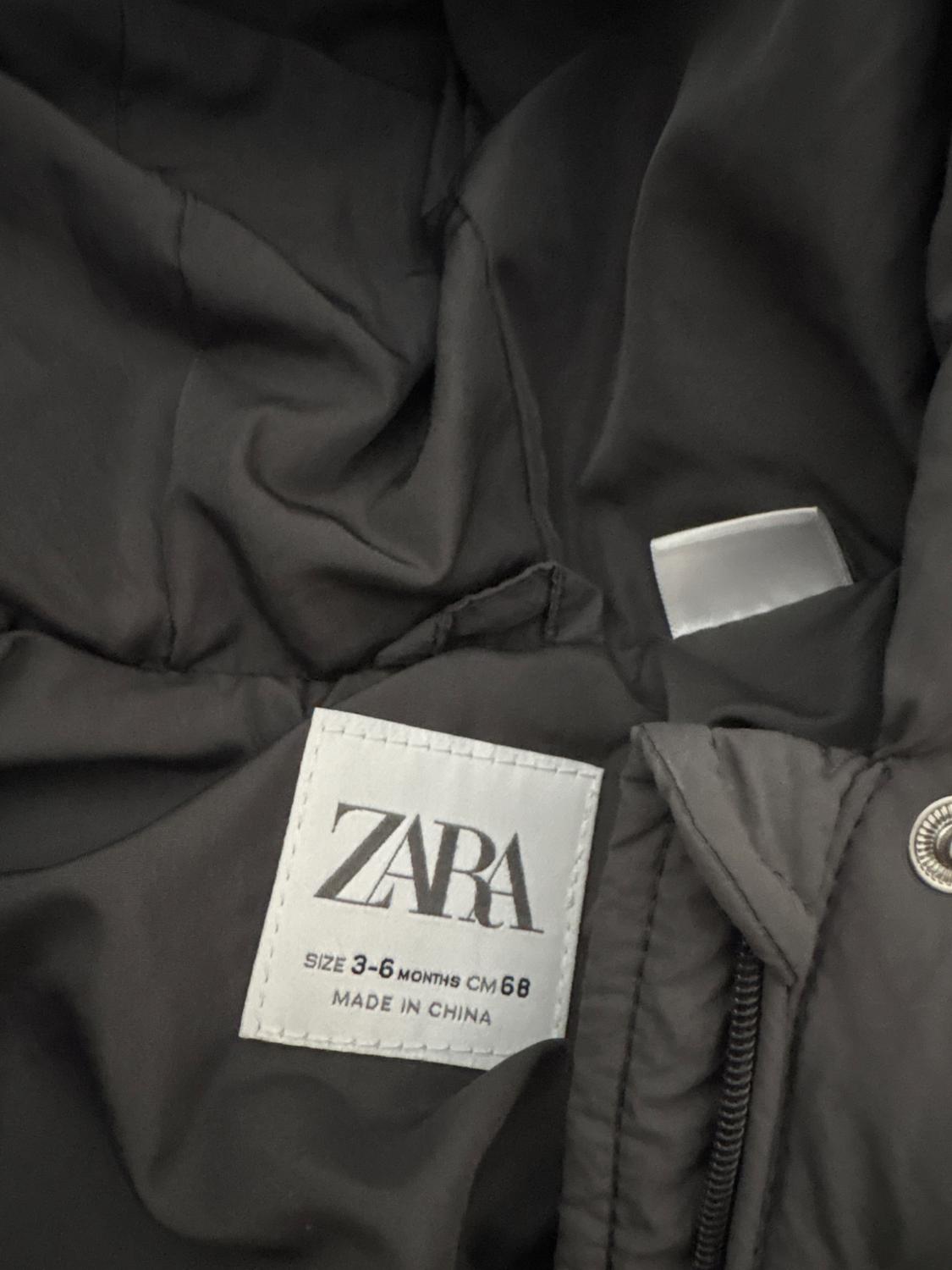 Zara white label on a black garment, indicates size 3-6 months, CM 68, made in China. Wrinkled fabric and visible metal button.
