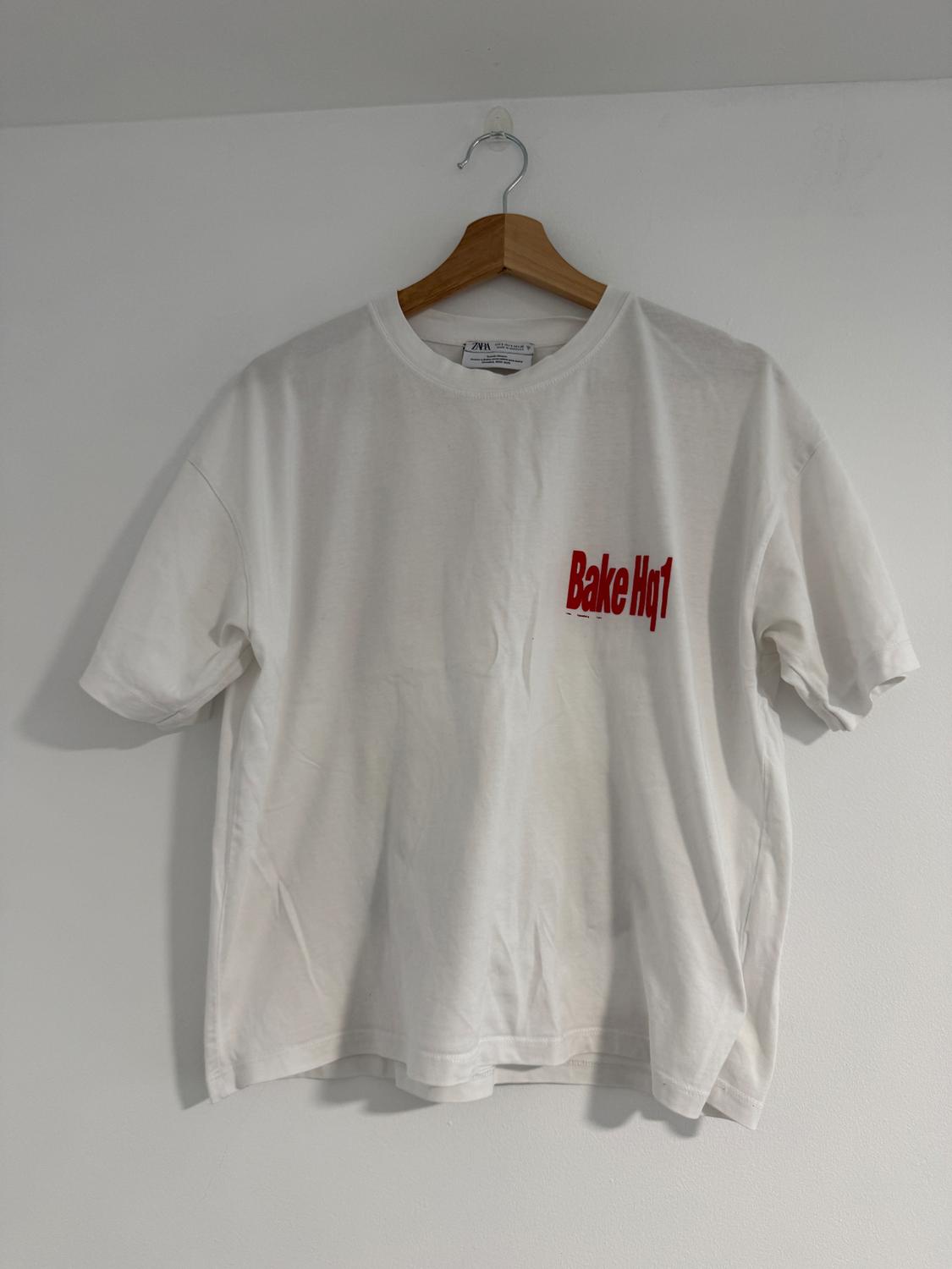 White t-shirt hung on a wooden hanger, with red text "Bake Hq1" on the chest. White background.