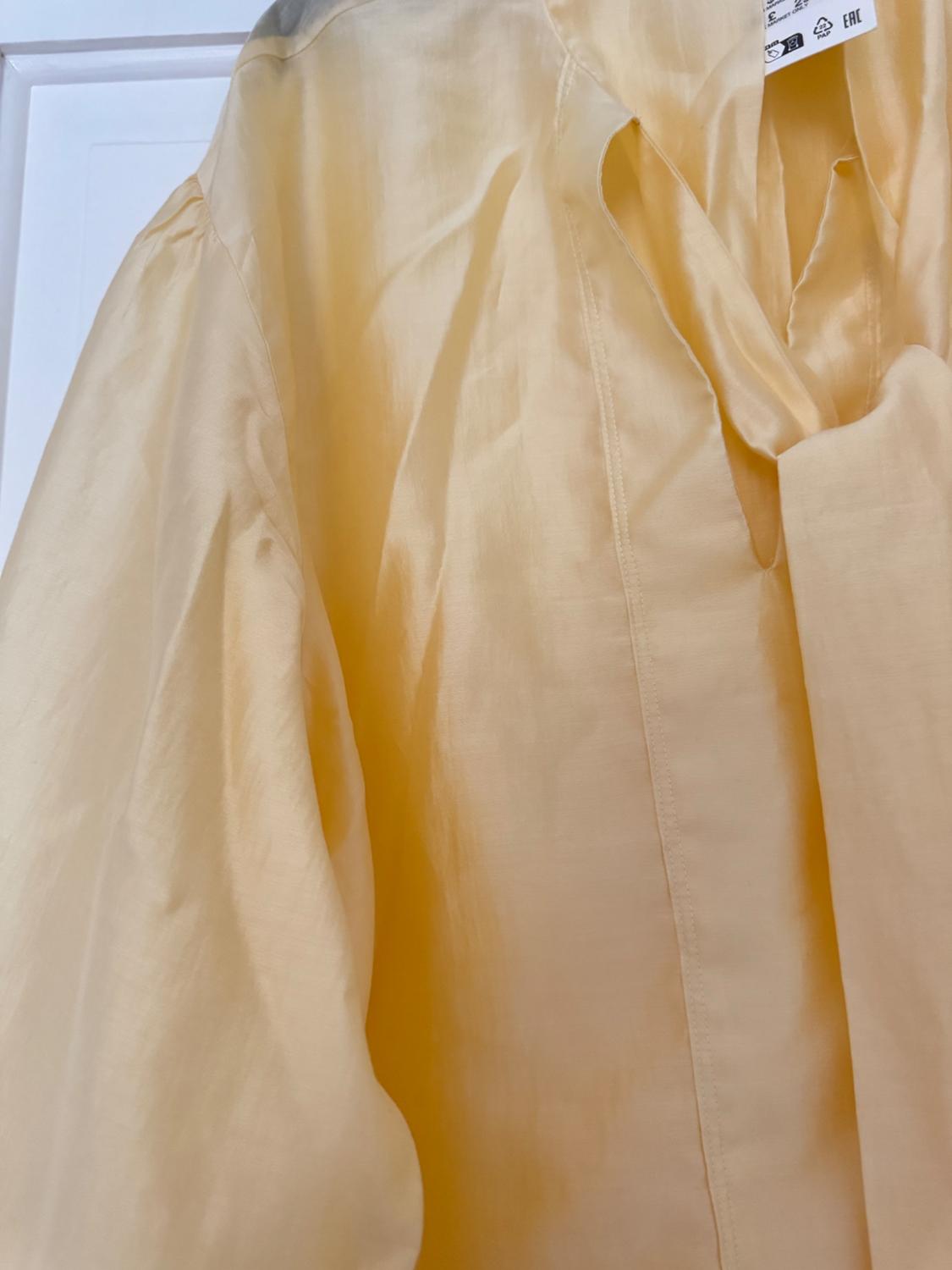 Close-up of a light yellow blouse, shiny fabric, V-neck and tie detail. Relaxed style. Label with text EAC PAP 22. White background.