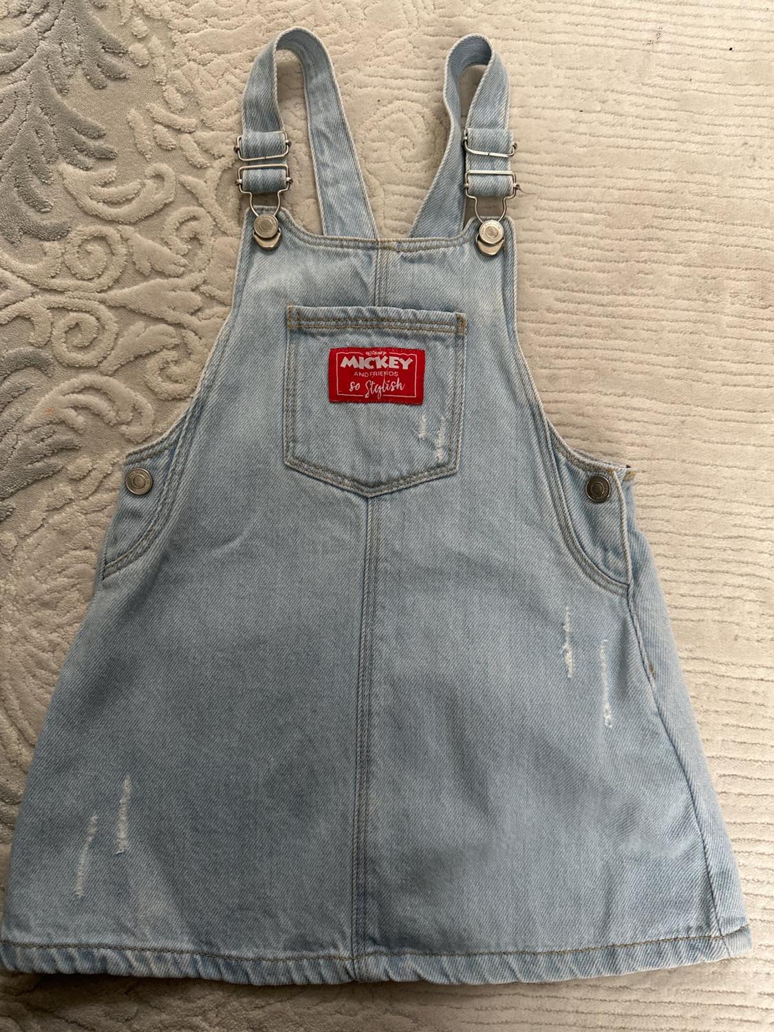 Front view of light blue denim pinafore dress with front pocket and text 'Mickey and Friends So Stylish'.