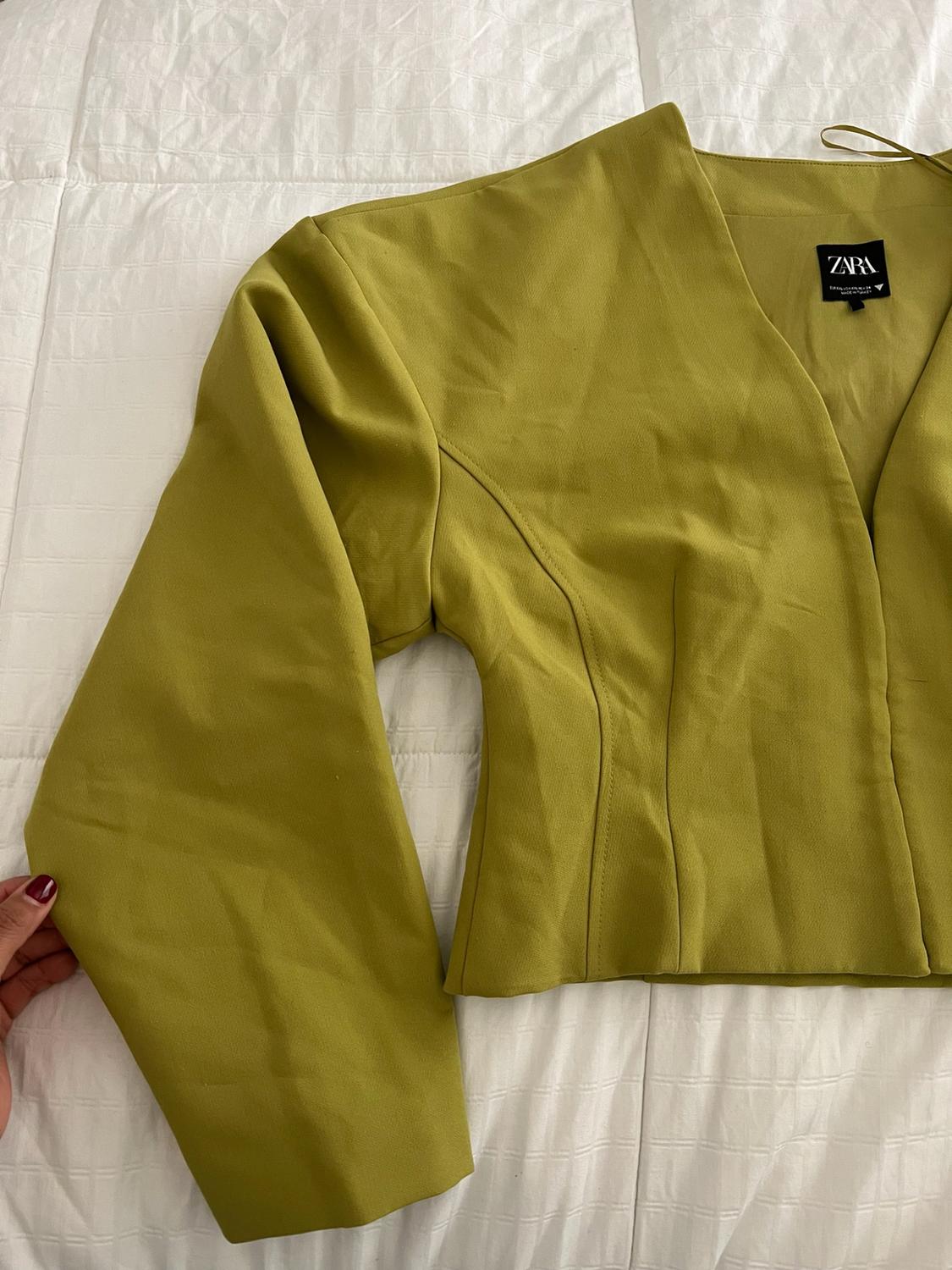 A green long-sleeve V-neck top rests on a white bed. Someone grabs the sleeve with their hand, and a label with the word ZARA can be seen.