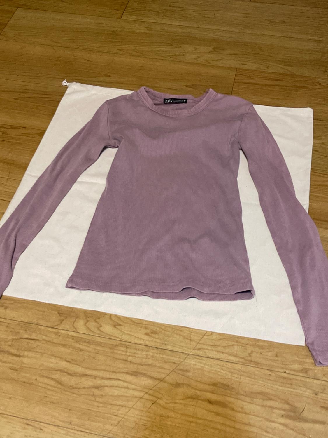 Lilac long-sleeve t-shirt on a white fabric on the wooden floor. Visible tag on the collar.