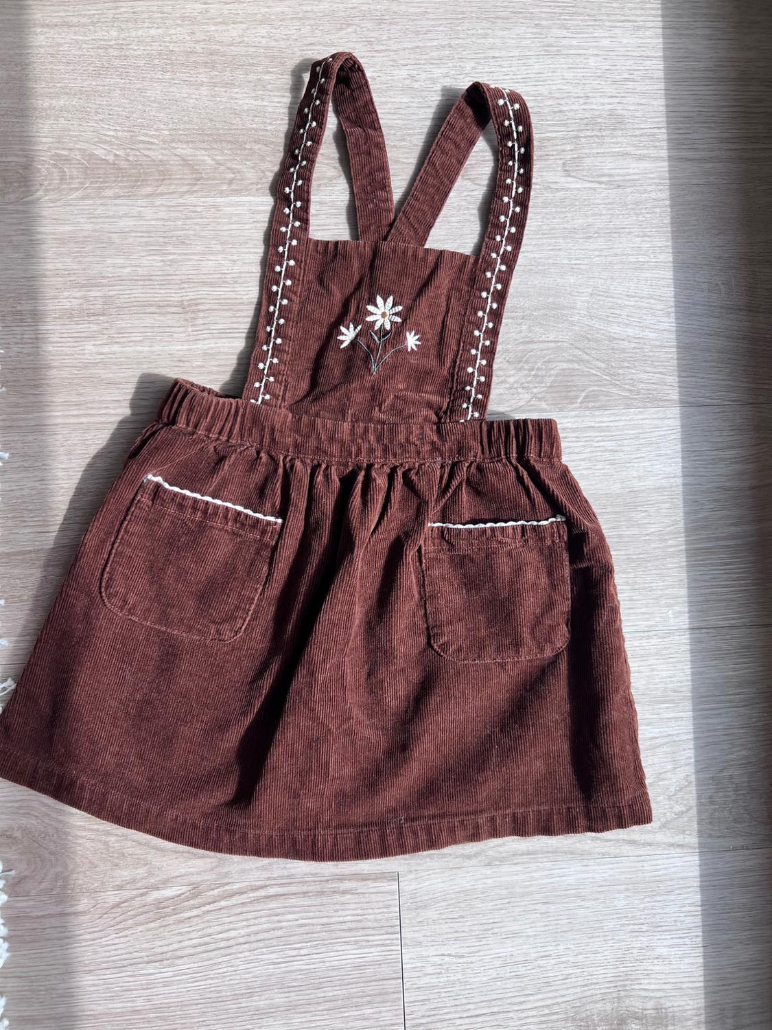 Brown corduroy kids’ jacket with white flower embroidery on the chest and two pockets with piping, on a light wood floor.