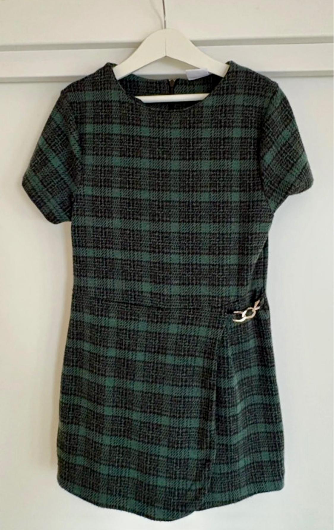 Green and black checked short dress with short sleeves, hanging on a white hanger. Gold brooch detail at the waist.