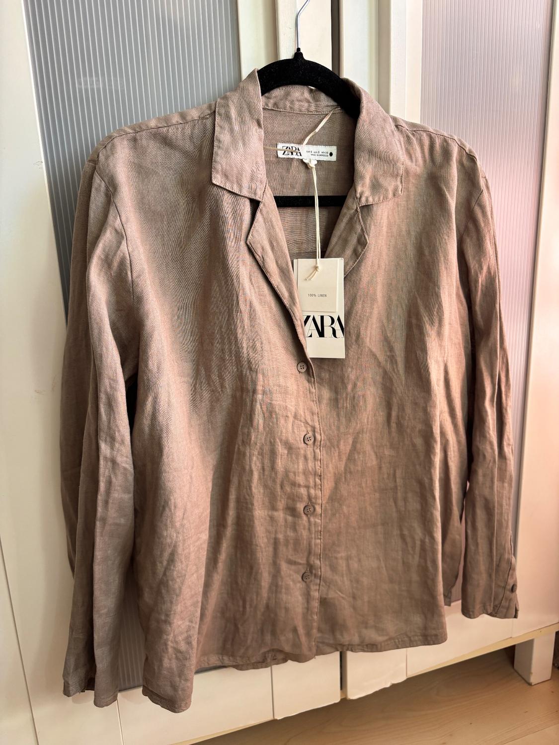 Beige linen shirt hanging on a black hanger. Visible label with text: "ZARA, 100% LINEN". White door background.
