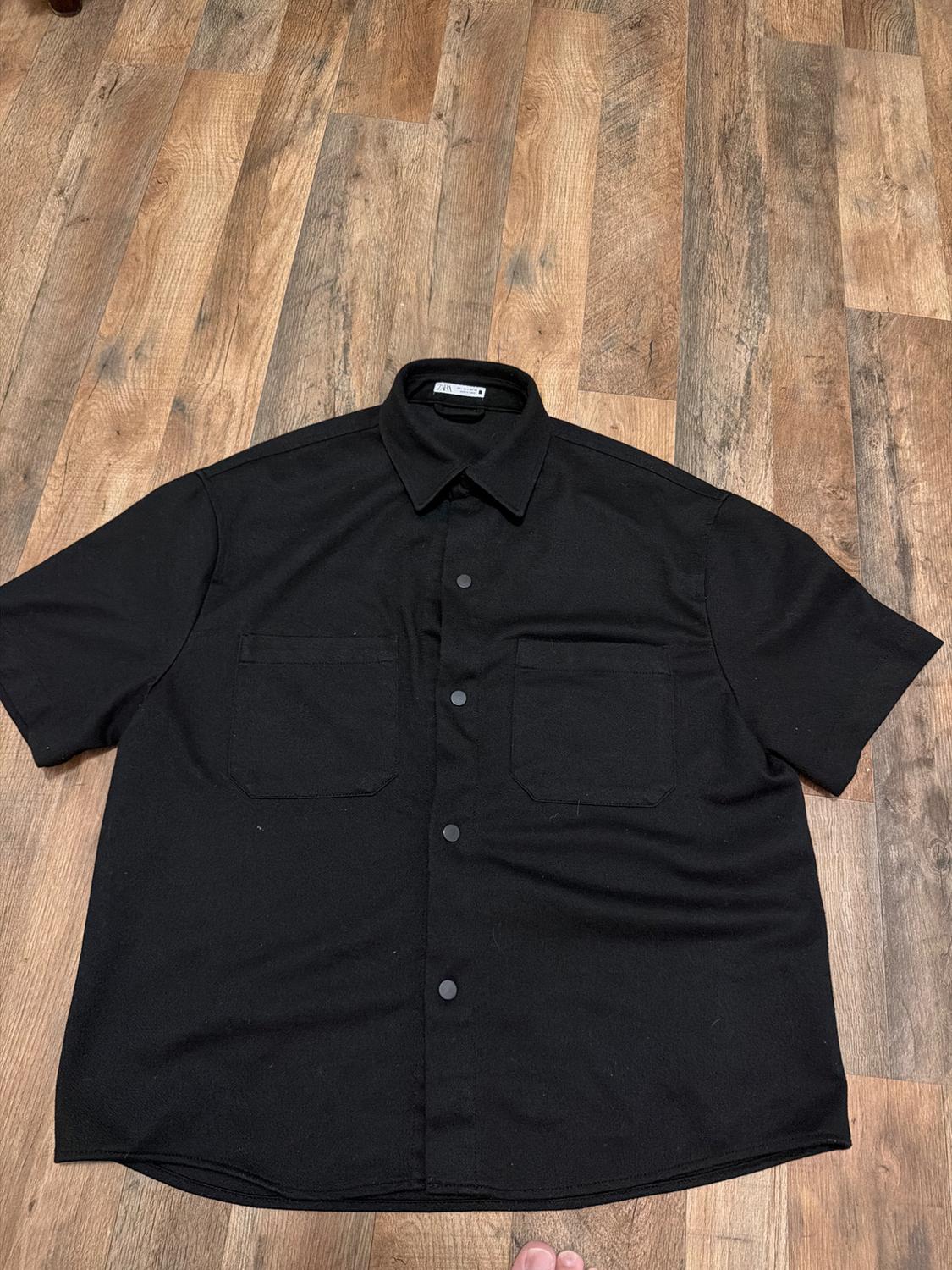 Short-sleeve black shirt with a collar, snap buttons, and two chest pockets, spread out on a wooden floor. Visible Zara tag.