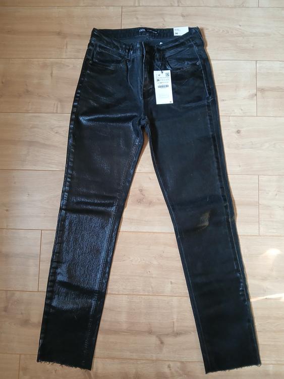 Shiny black trousers on a wooden floor, with a visible price tag. Slim fit and frayed hem.