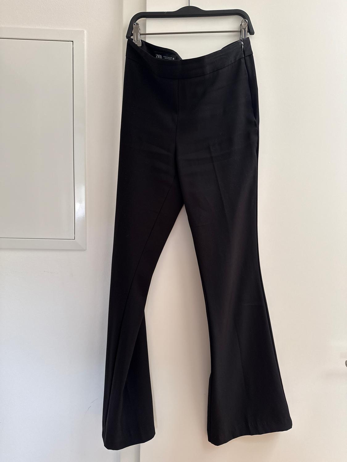 Black flared trousers hanging on a black hanger over a white door. Label visible on the waistband.