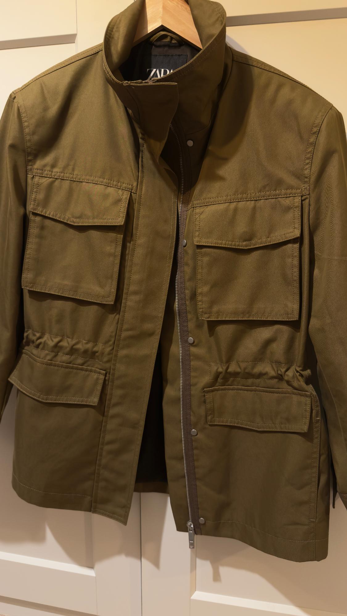 Olive green jacket with large pockets and zip, hanging on a wooden hanger. Visible label with text "ZARA".