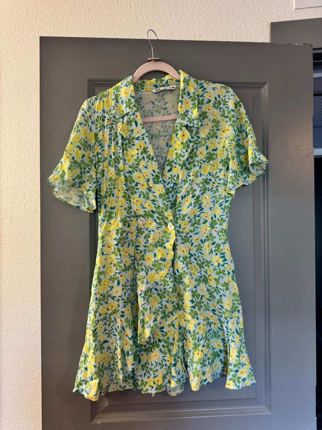 Short dress with a floral print in shades of green and yellow, hanging on a hanger over a grey door.