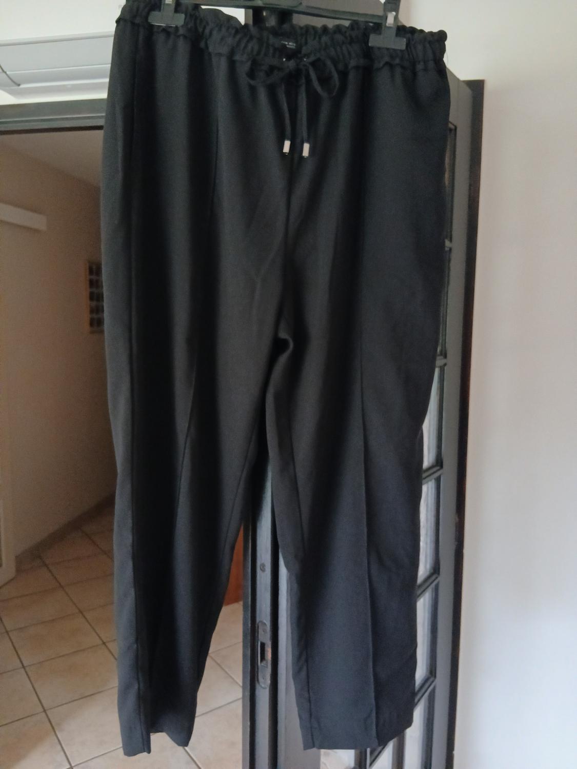 Black trousers hanging on a glass door, with an elasticated waist and adjustable drawstring, in an indoor setting with a tiled floor.