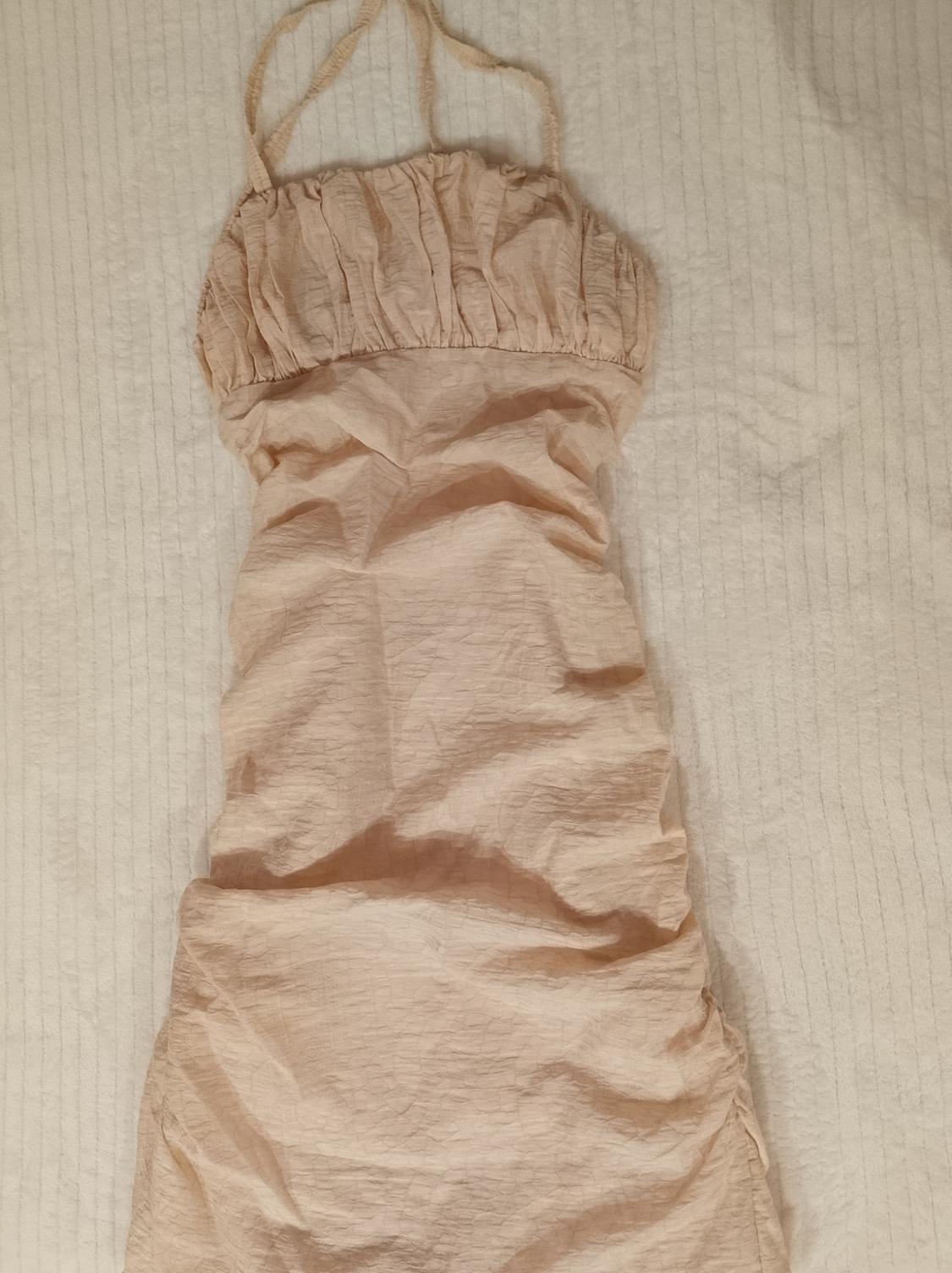 Light beige slim-fit dress with thin straps and textured fabric with ruching, laid out on white fabric. Relaxed composition.