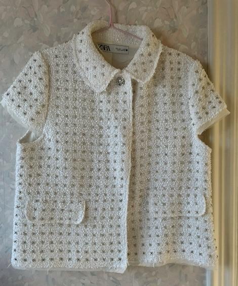 White knitted jacket with a small check texture and short sleeves, hanging on a hanger against a floral background.