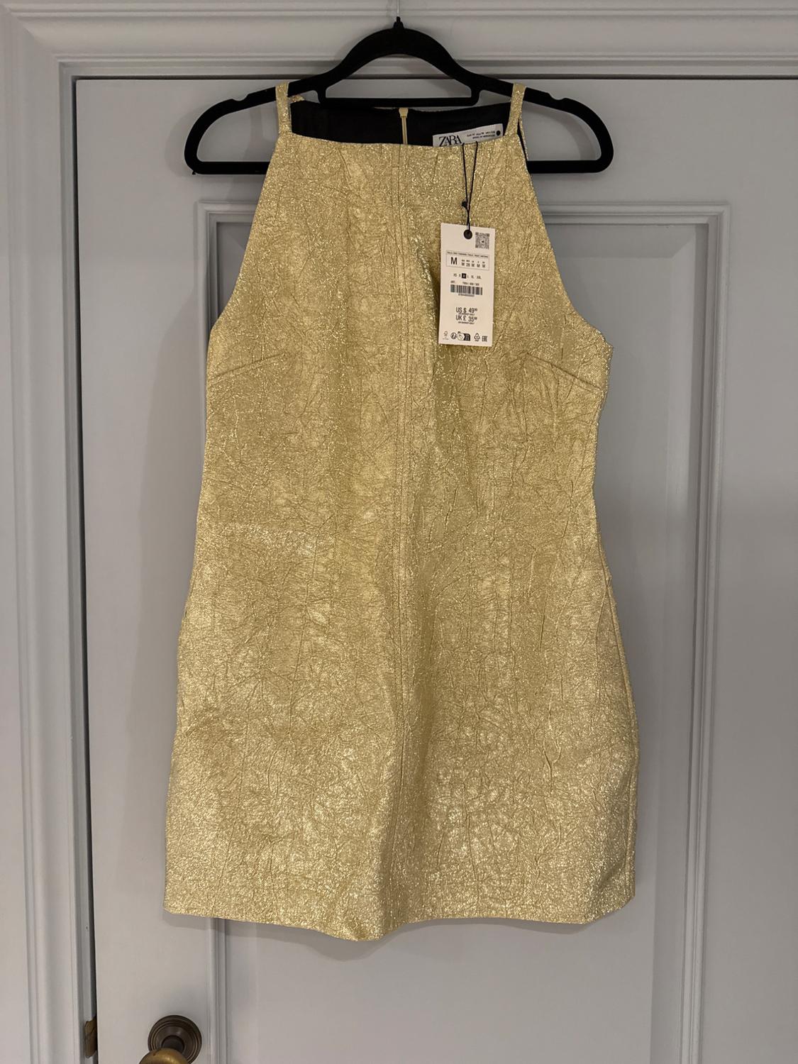 Sleeveless gold dress hanging on a black hanger over a grey door. Visible tag with price and size M.