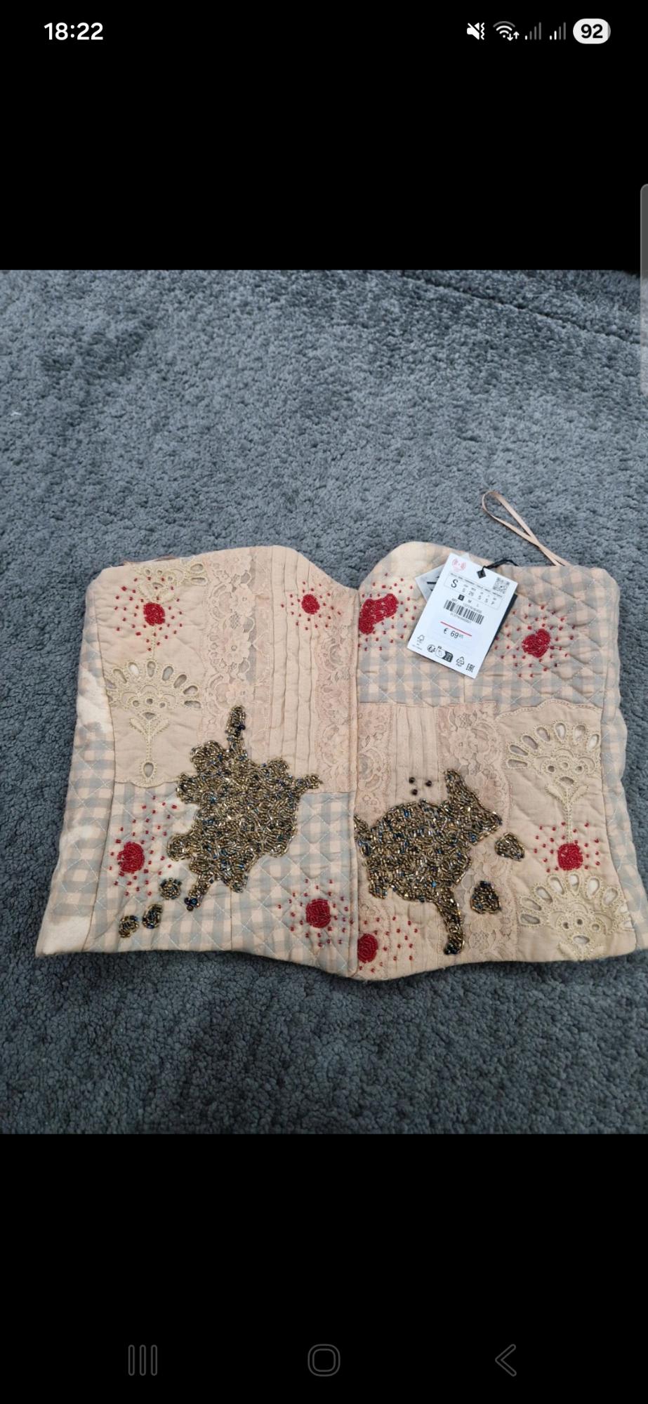 Beige patchwork top on a grey rug with lace, beads and embroidery. Label with text S € 69.95.