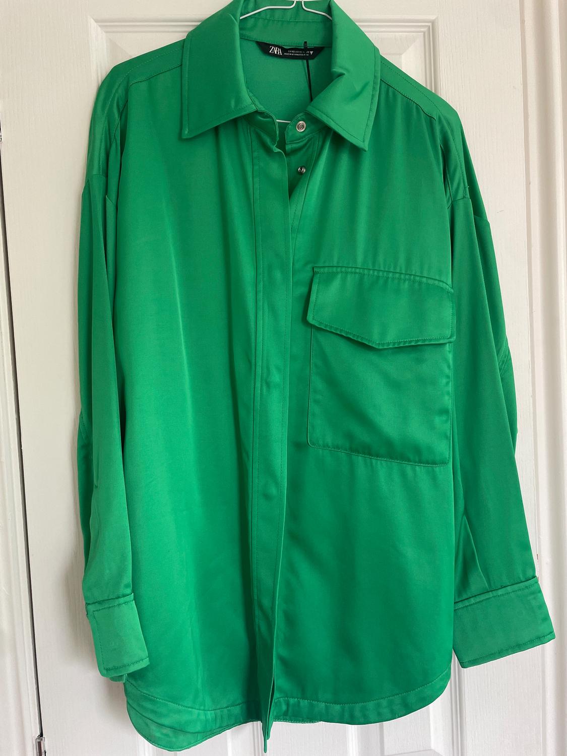 Green shirt hanging on a white hanger, with a front pocket and classic collar. Visible label inside the collar.