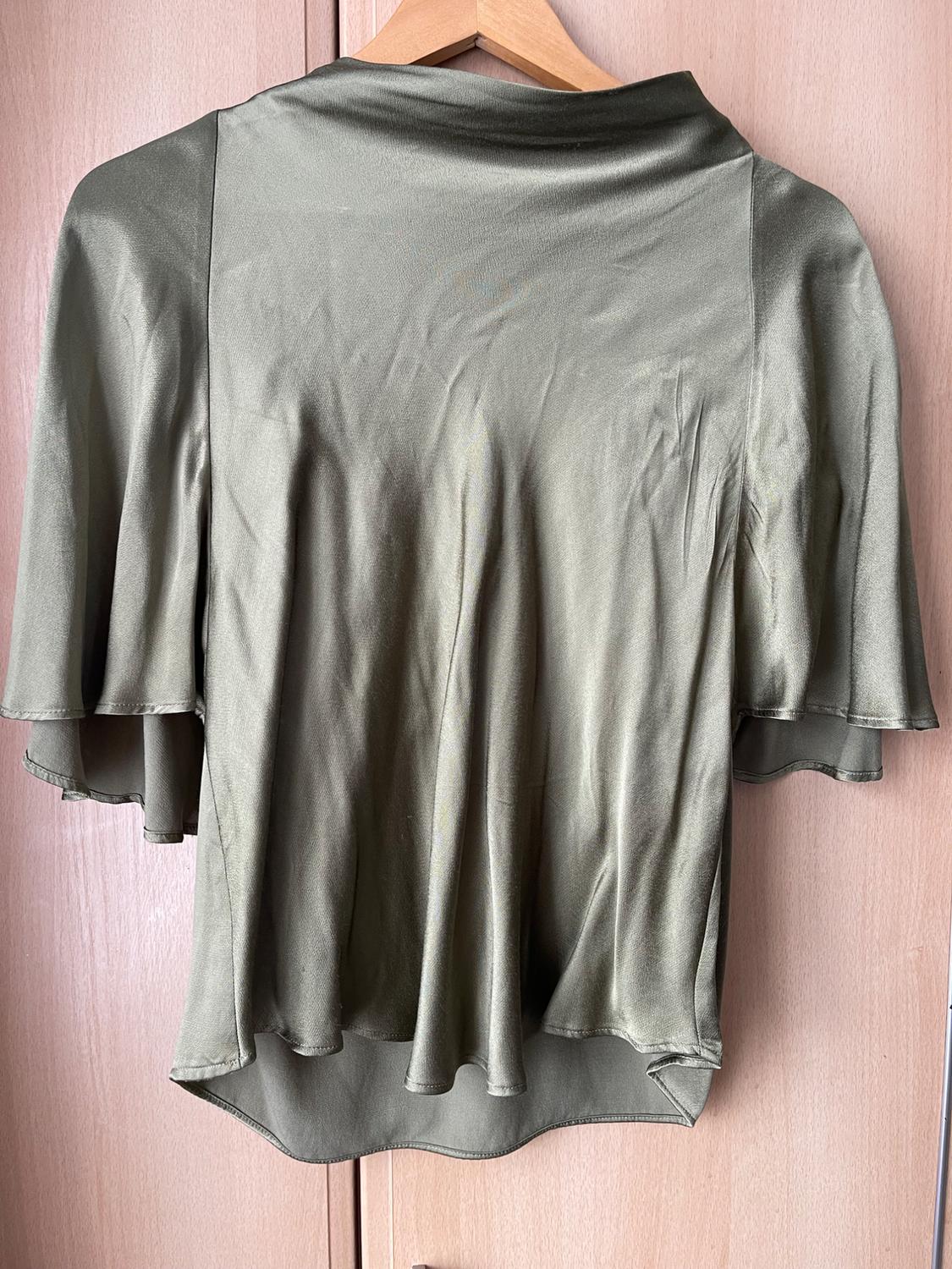 Olive green satin blouse with voluminous sleeves, hanging on a wooden hanger against a light wood wardrobe background.