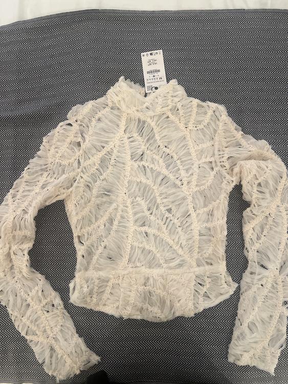 White lace blouse with leaf design, long sleeves, over grey herringbone fabric. Visible label on the collar.