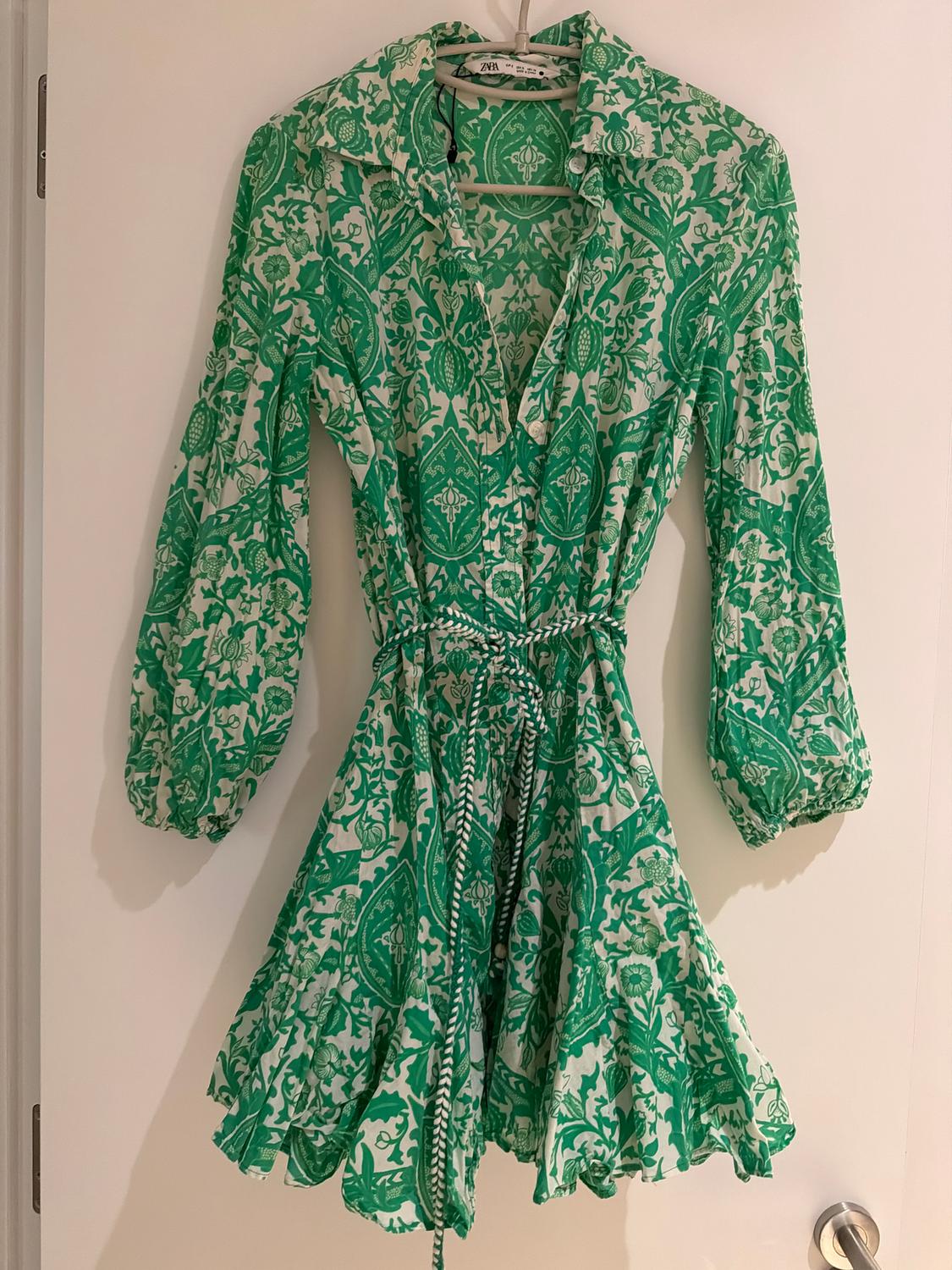 Short green dress with a white floral print, puff long sleeves and a braided belt, hanging on a white hanger.