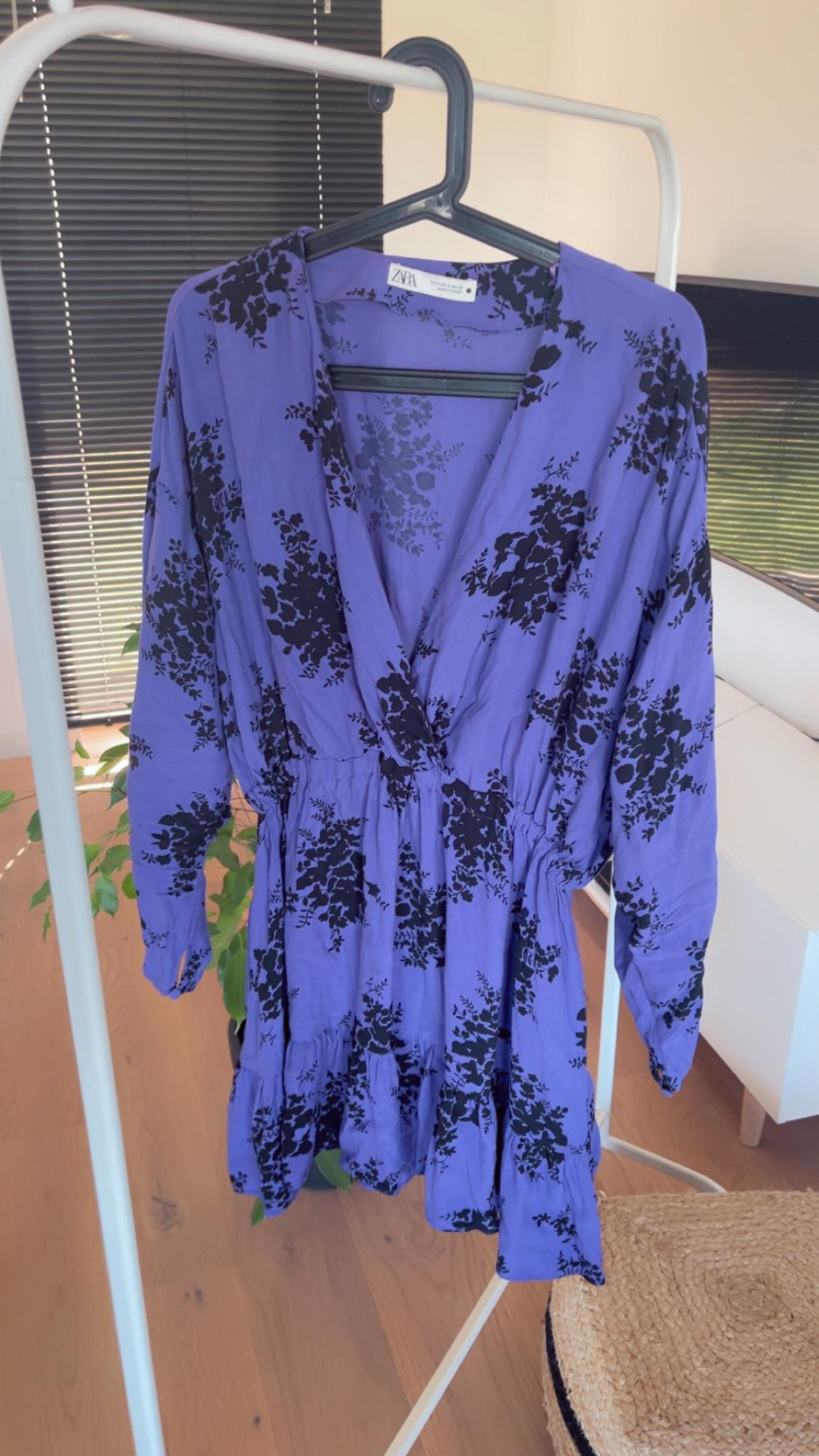 Purple dress with a black floral print hanging on a white clothes rail, in a room with a wooden floor and a decorative plant.