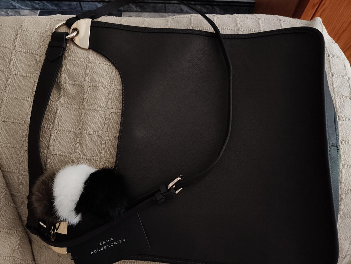 Black handbag with a decorative pom-pom on a textured beige surface. Includes a tag bearing the text “ZARA ACCESSORIES”.