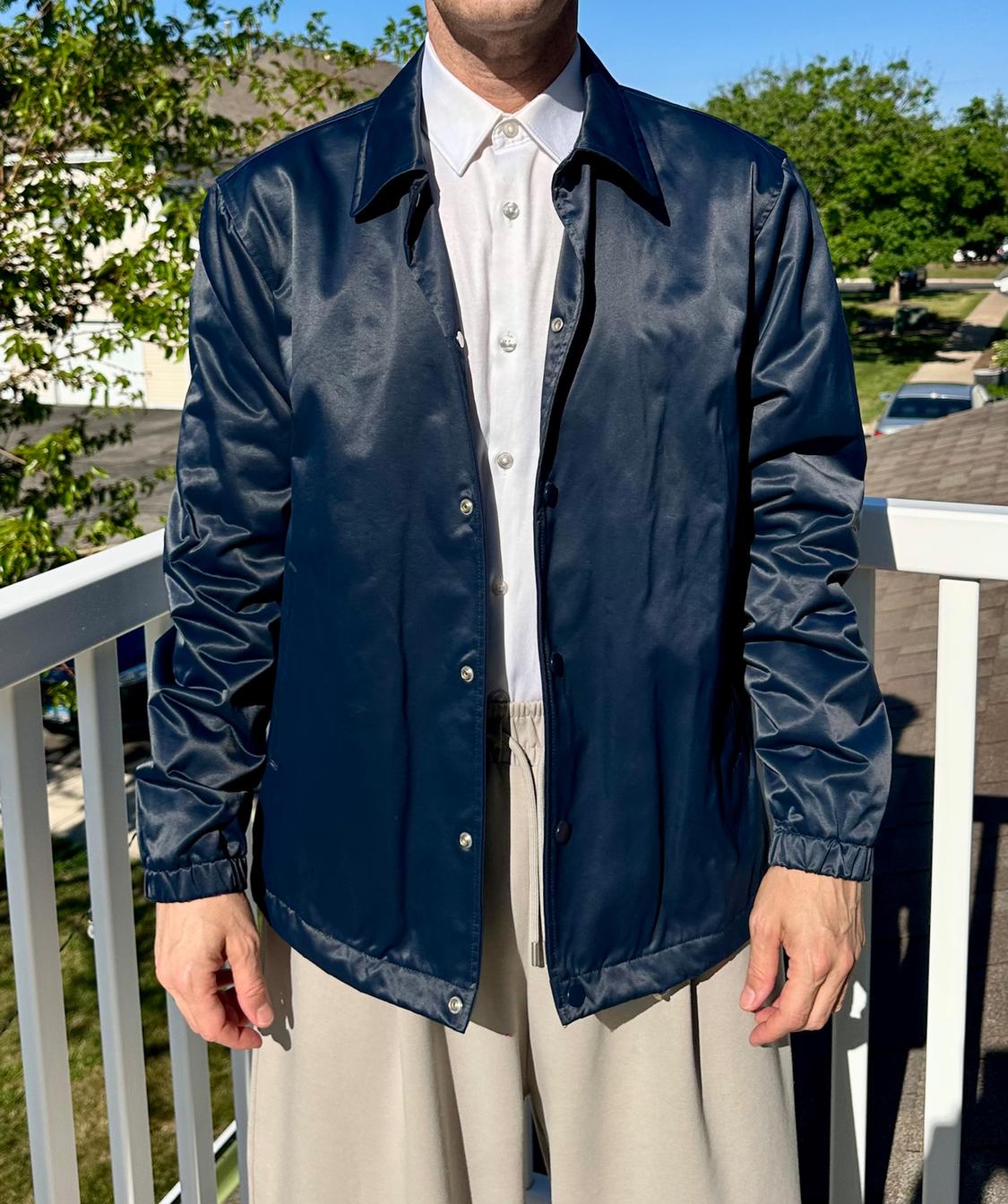 Man in a dark blue jacket and white shirt on a sunny balcony. He's wearing beige trousers. Background of trees and houses.