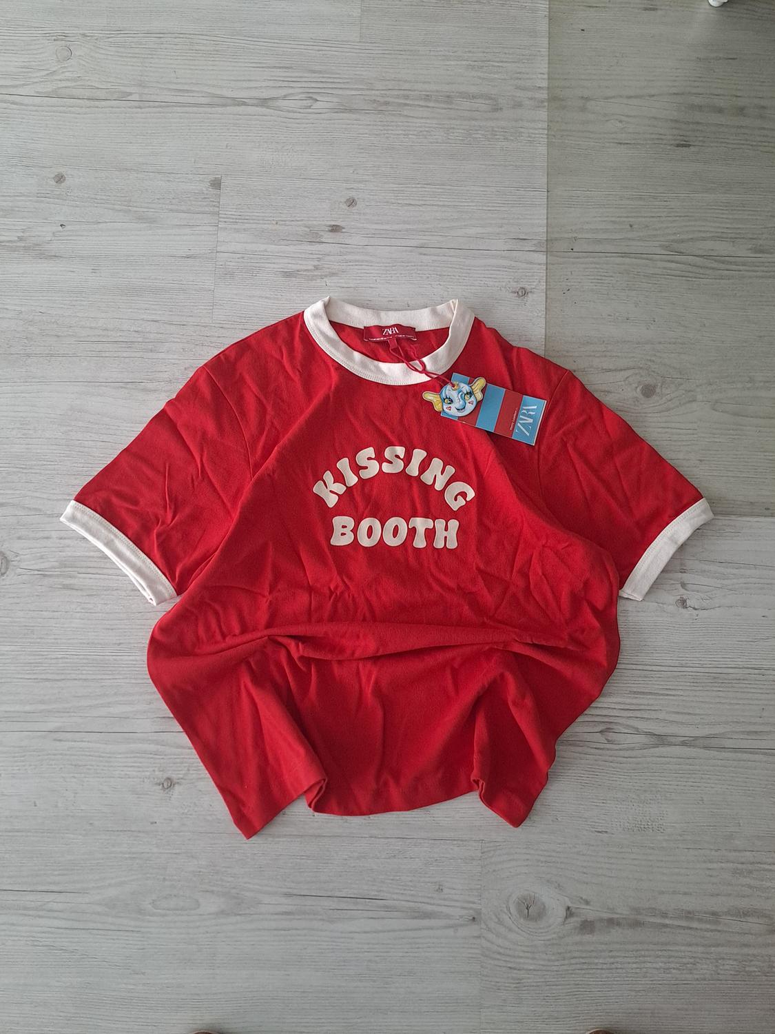 Red t-shirt with white collar and sleeves on a light wooden floor. Text "KISSING BOOTH" on the front. Hanging blue tag.