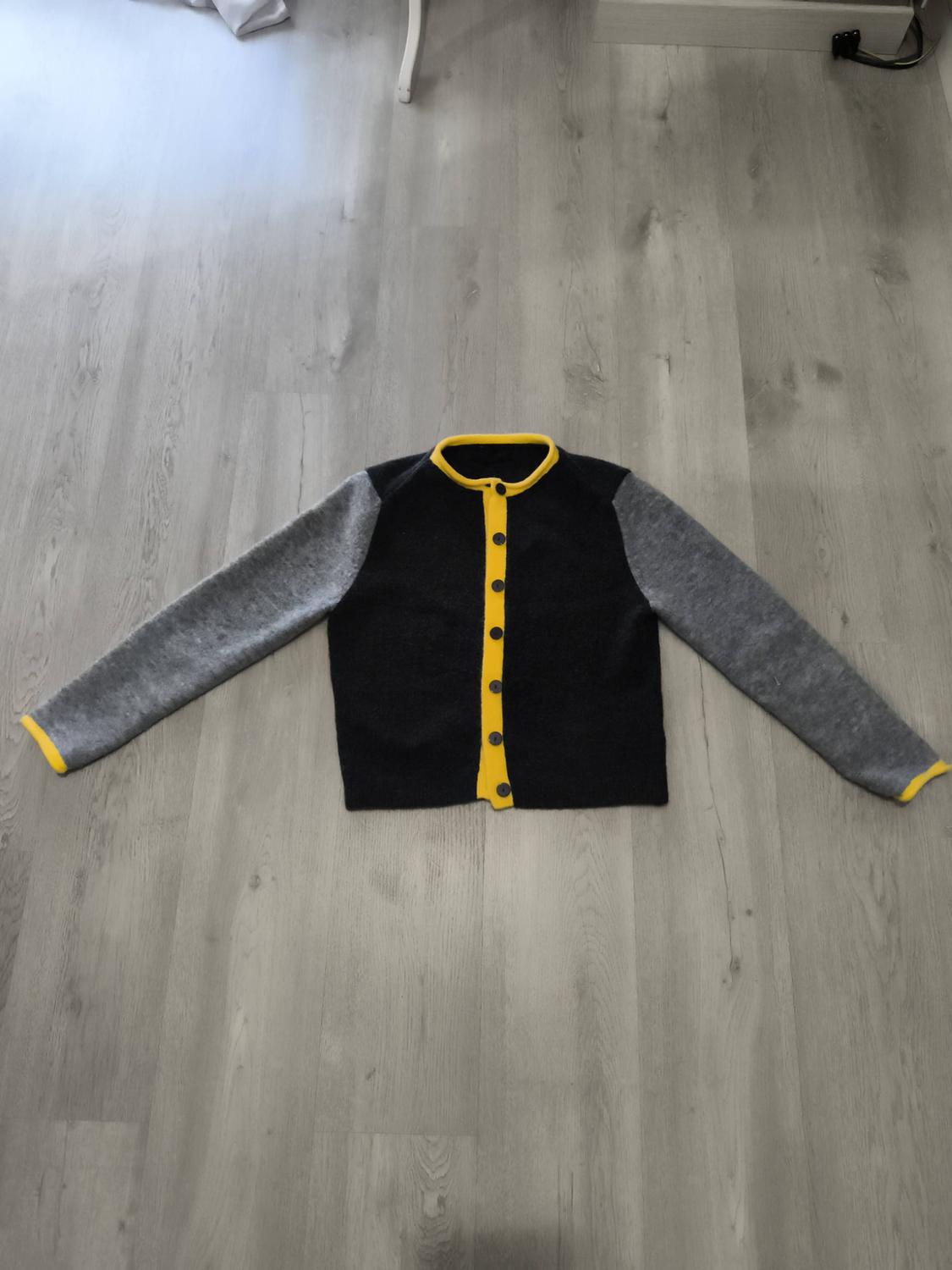Wool jacket on the floor, black body with buttons and a yellow collar, grey sleeves with yellow cuffs, on a light wooden floor.
