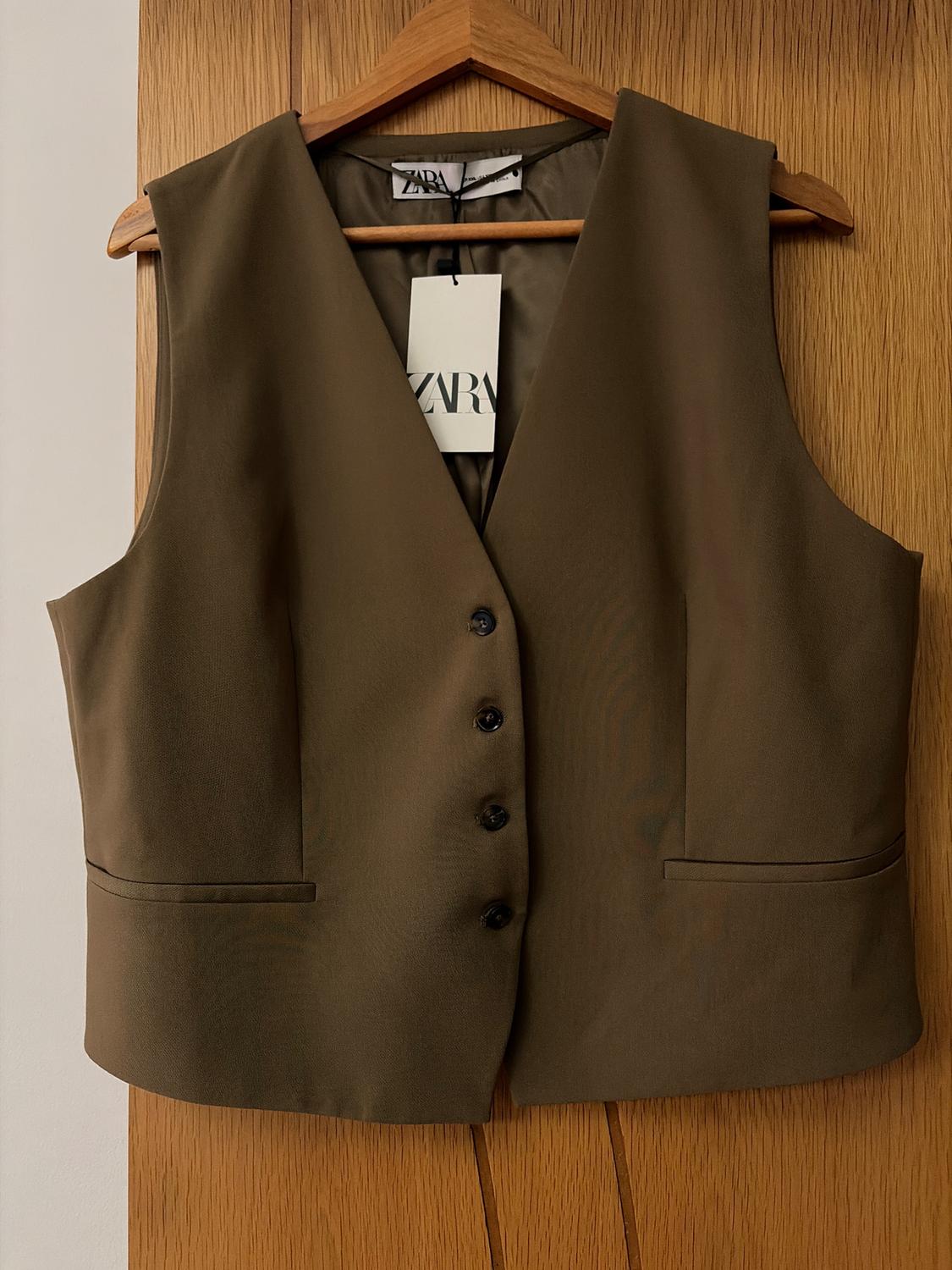 Brown sleeveless waistcoat with black buttons, hanging on a wooden hanger. Visible label with the text "ZARA". Light wood background.
