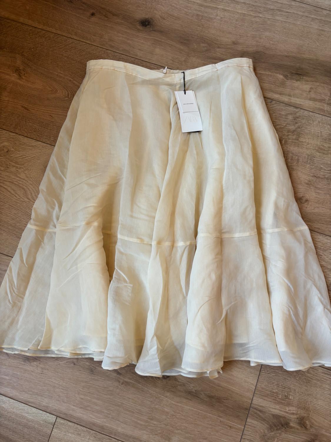 Cream-coloured flowing midi skirt with horizontal seams, laid out on a wooden floor. ZARA label visible.