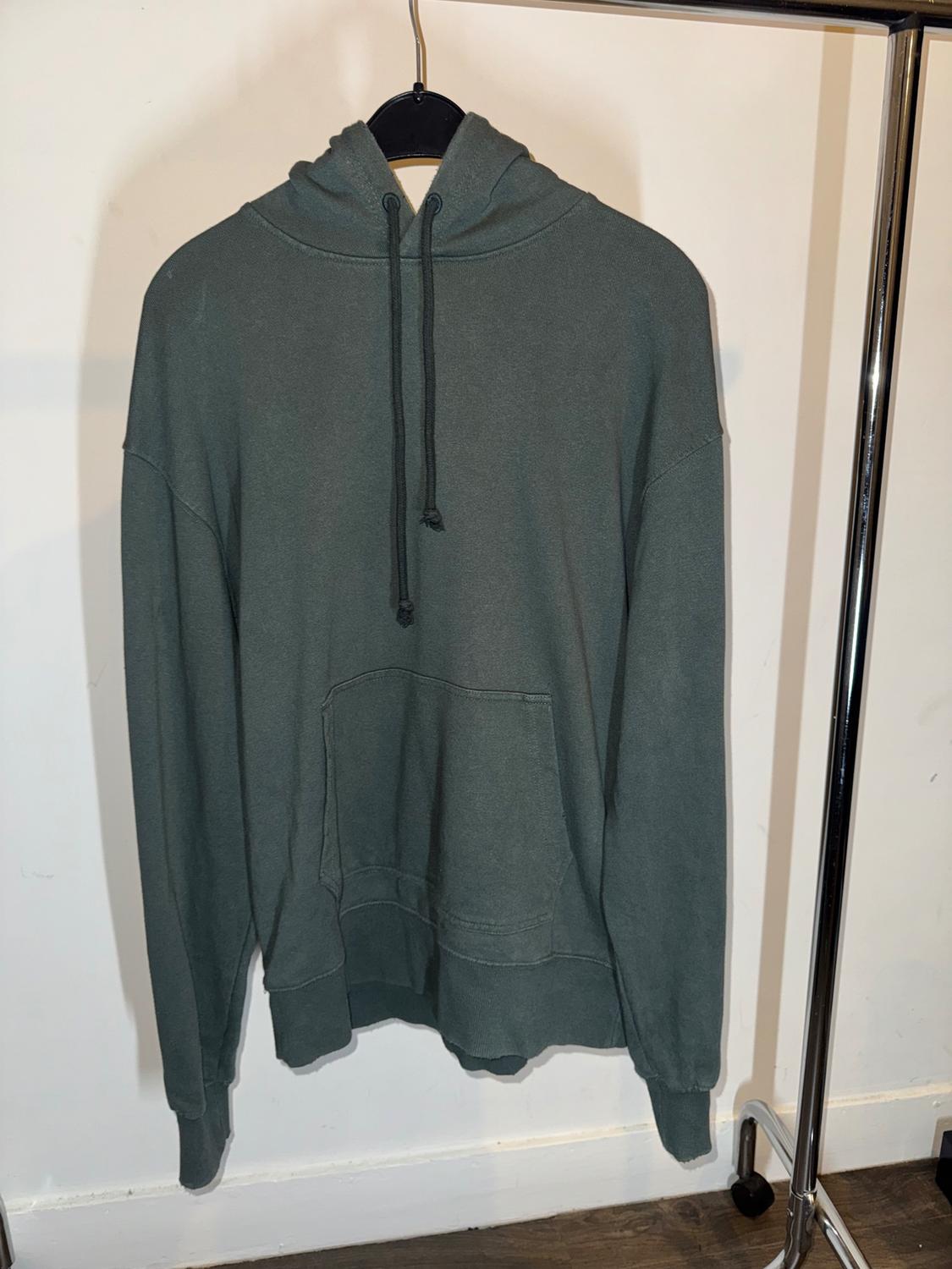 Dark green hoodie hanging on a black hanger against a white background. It has a front pocket and adjustable drawstrings.