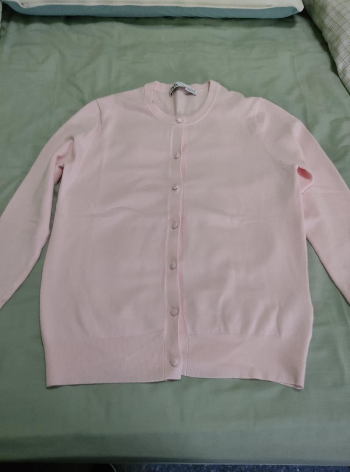 Light pink crew-neck, long-sleeved cardigan with front buttons, laid out on a green sheet. Casual and relaxed style.