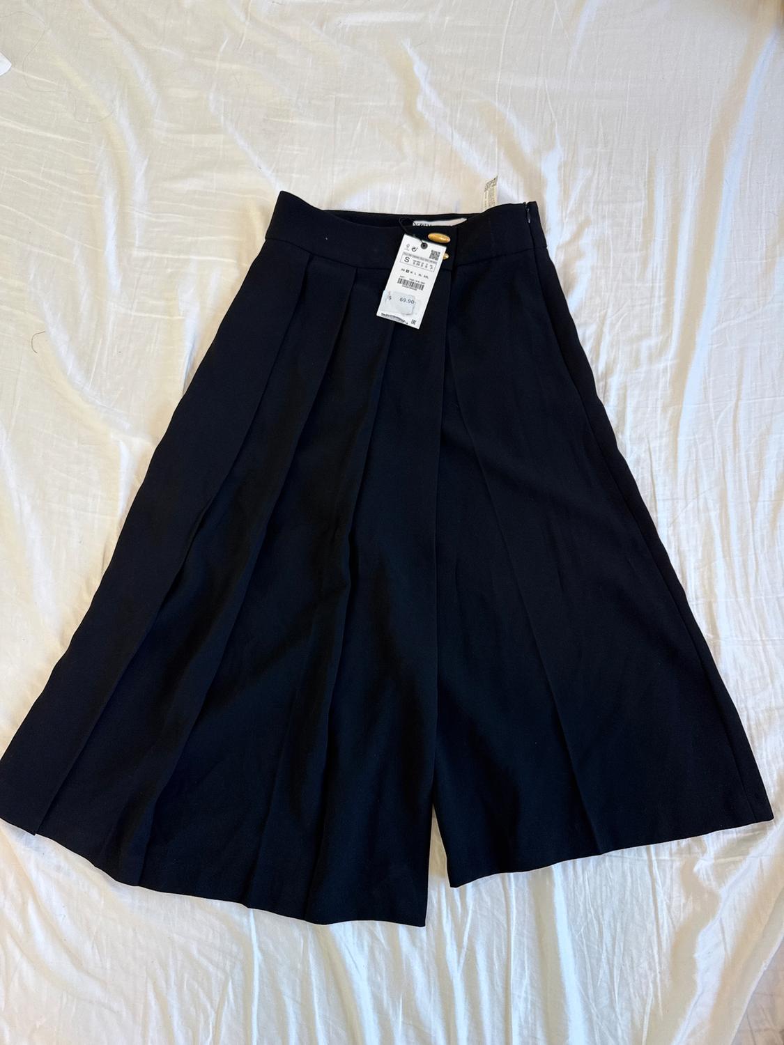 Black shorts with pleats on a white sheet. Visible tag with price and size.