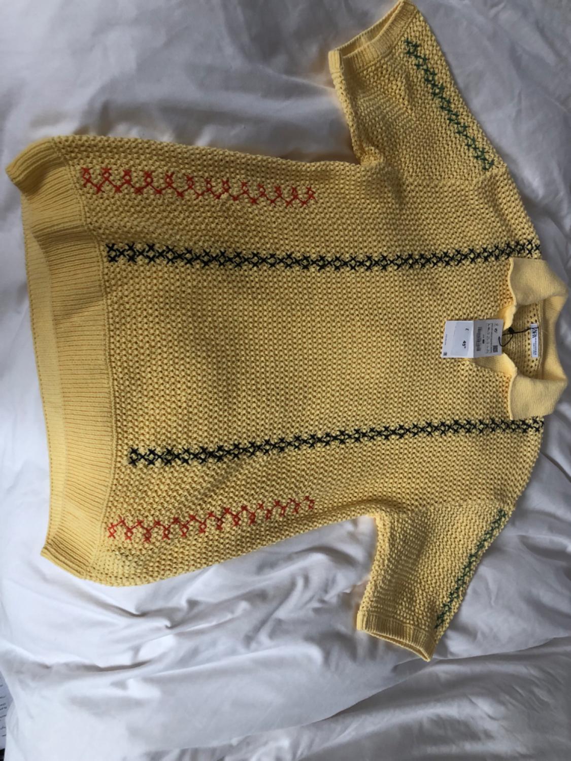 Yellow chunky knit jumper with stitching details in red, black, and green, on a white bed. Visible neck label.