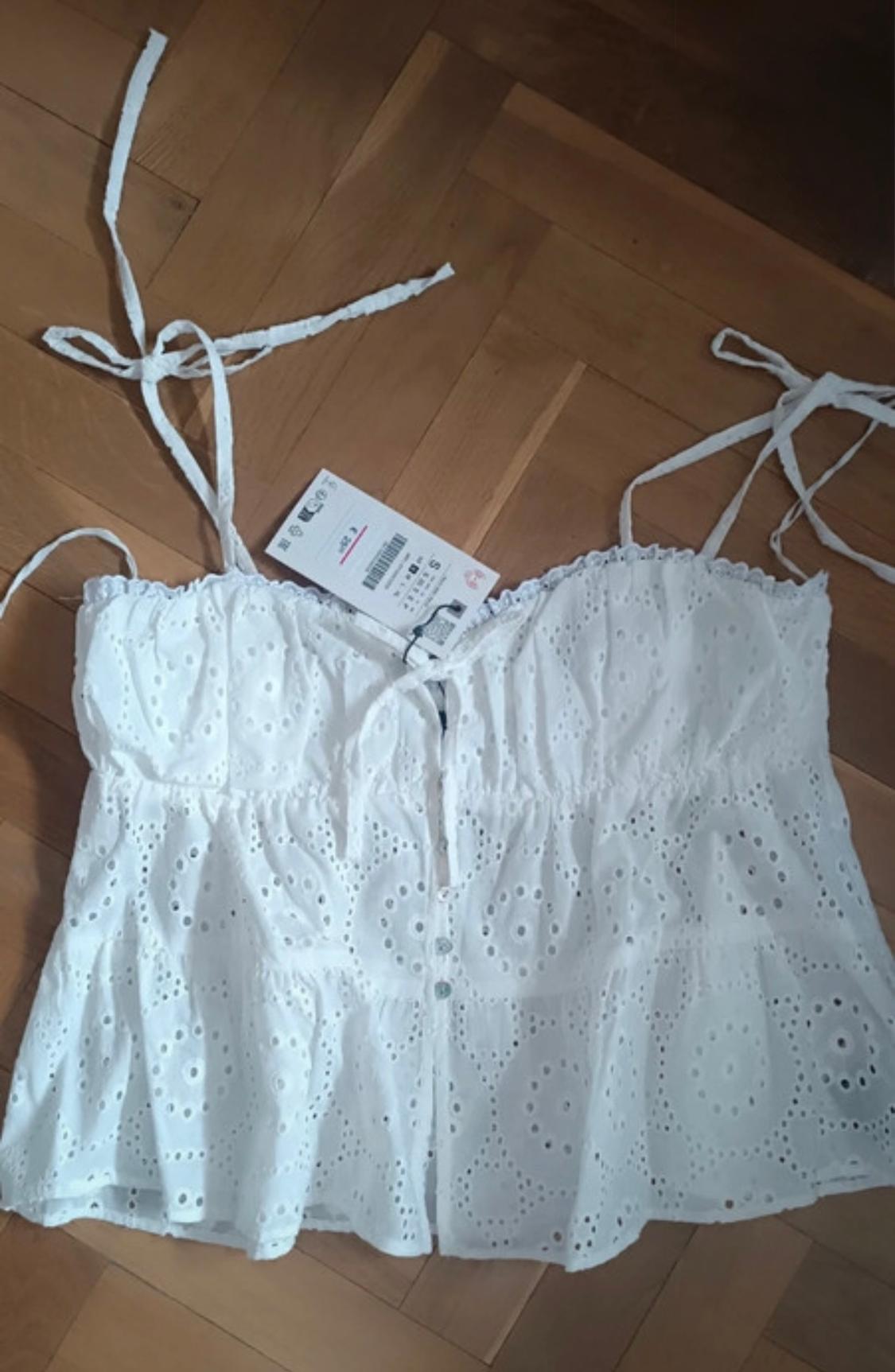 White sleeveless top with openwork embroidery on a wooden floor. Visible label with text in another language.