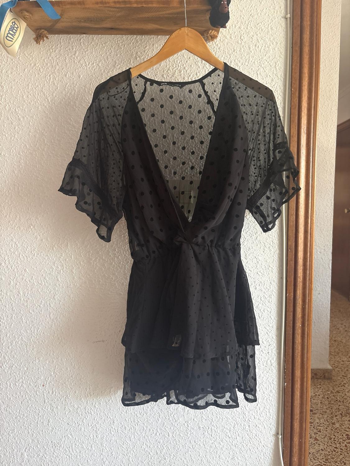 Black semi-sheer polka dot V-neck blouse with frilled sleeves and a front tie, hanging on a hanger against a white wall.