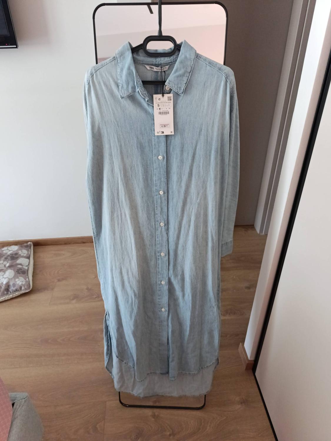 Light blue denim shirt dress with front buttons, hanging on a black clothes rack in a room with a wooden floor.