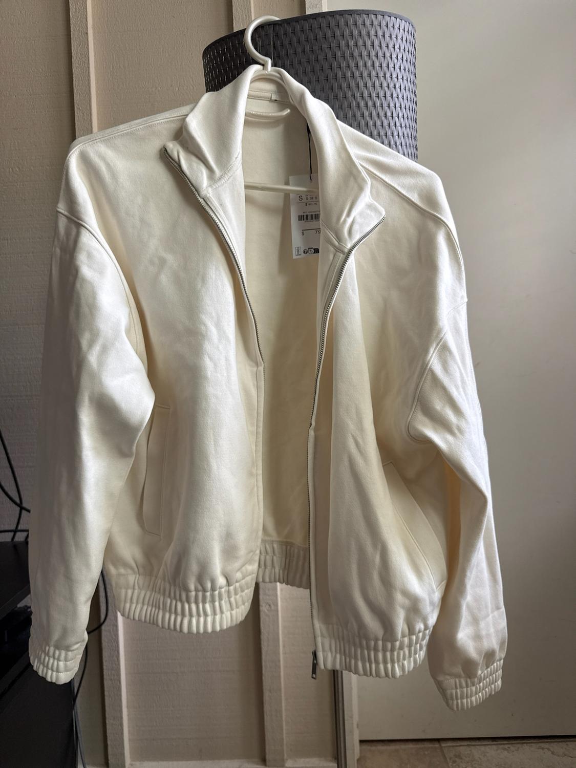 Cream-coloured jacket with a zip, high collar and visible label, hanging on a coat hanger indoors.