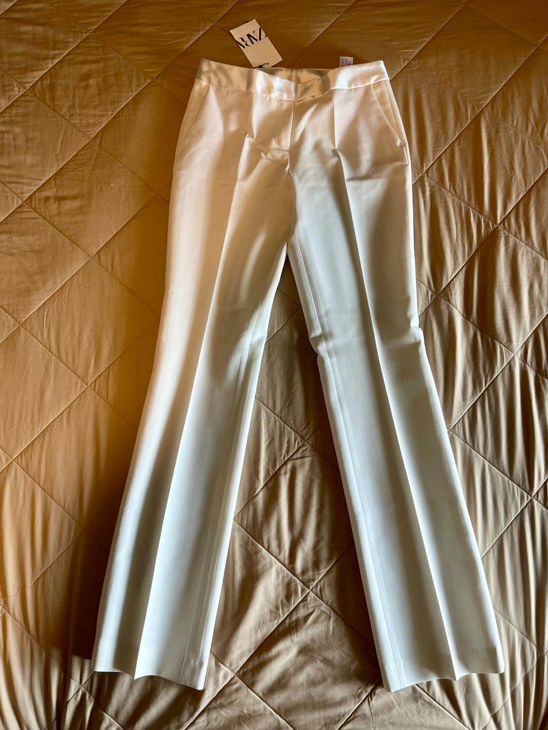 White pants with a front crease and ZARA tag, spread out on a beige quilted bedspread.