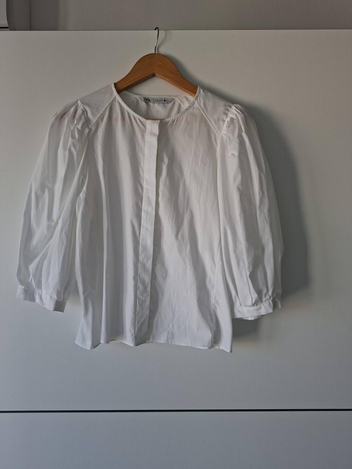 White round-neck blouse with three-quarter-length puff sleeves, hanging on a wooden hanger against a white background.
