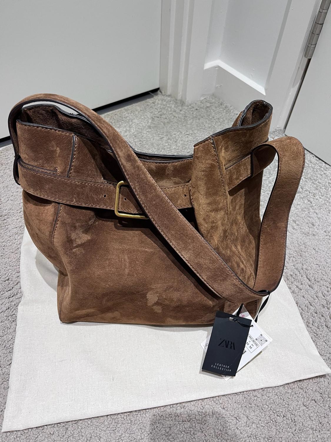 Brown suede bag with a strap and buckle over a white cloth on the floor. Label with the text "ZARA LEATHER COLLECTION".