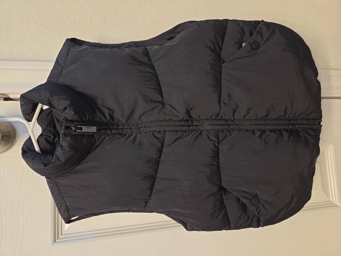 Black padded gilet hanging on a white over-door hanger. Sleeveless design with a front zip fastening.
