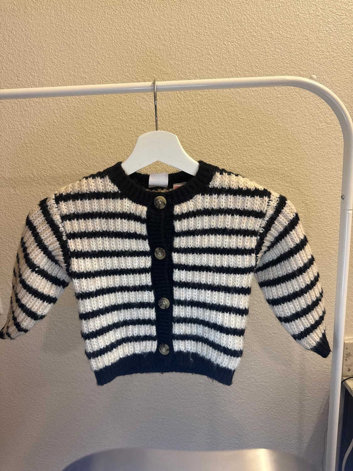 Black and white striped jumper hanging on a white hanger. It has large buttons on the front. Beige background.