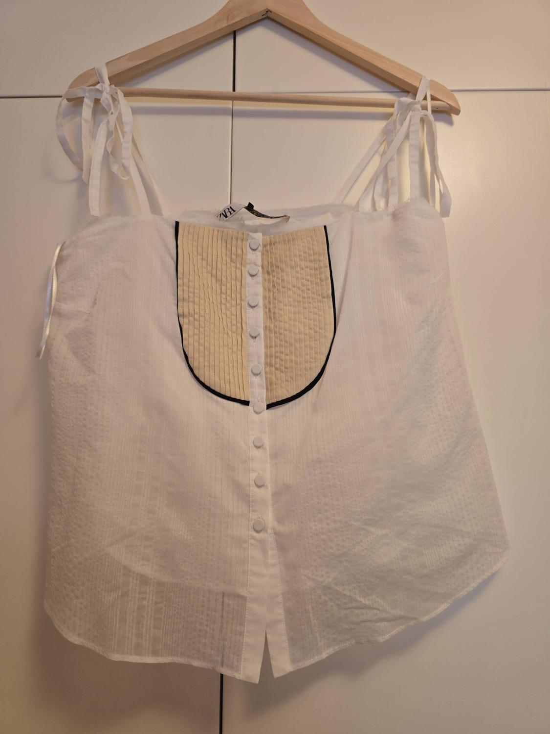 White strappy top with ties, buttons and a beige panel with black trim, hanging on a wooden hanger. ZARA label.