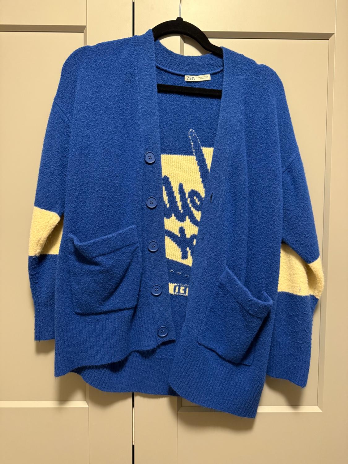 Blue knitted cardigan with buttons, pockets, yellow details, and lettering, hanging on a hanger in front of a light-colored wardrobe. Text: ZARA.