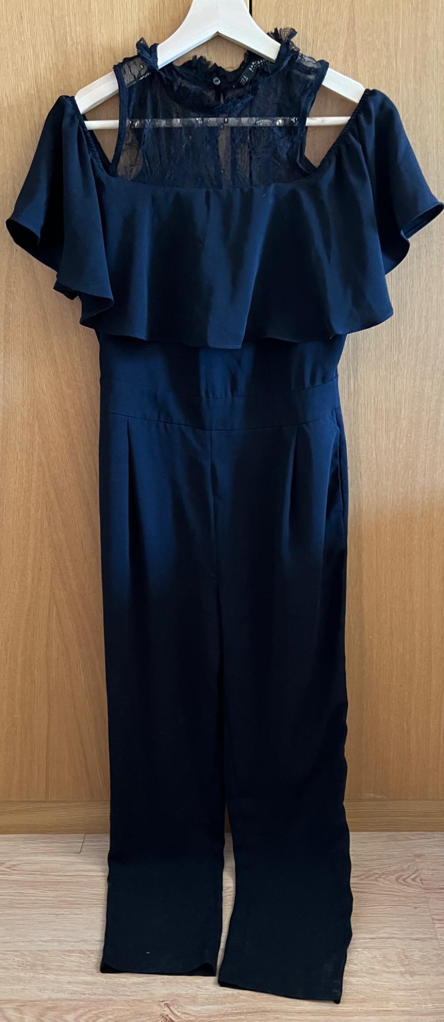 Navy blue full-length jumpsuit hanging on a white hanger against a wooden background. Notable features include its lace bodice, ruffles, and pleated trousers.