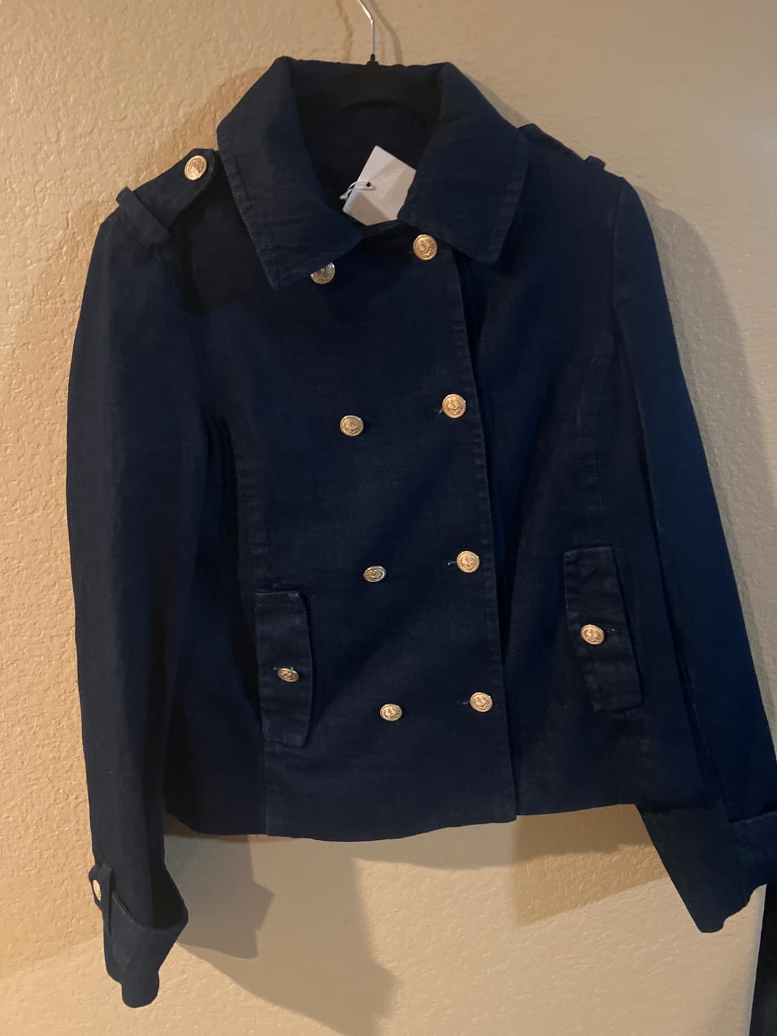 Dark blue jacket with gold buttons, hanging on a hanger against a beige wall. Double-breasted design with lapels.