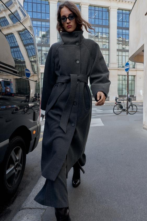 Dlouhý šedý kabát s vysokým límcem, dlouhými rukávy, předními kapsami a ladícím páskem.