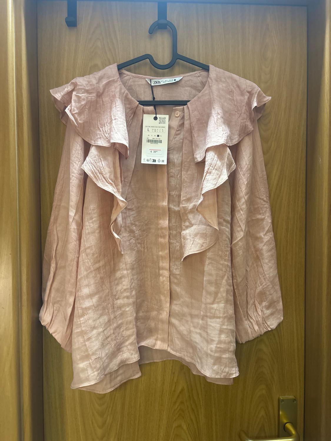 Light pink ruffled blouse hanging on a black hanger over a wooden door. Visible tag with price and size details.